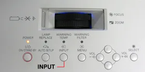 Bàn tay đang nhấn nút Input trên bảng điều khiển của máy chiếu Panasonic để chọn nguồn vào